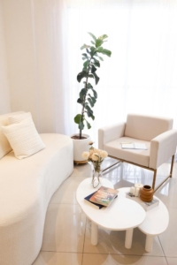 Bright, minimalist living room with a soft ivory sofa, white armchair, and a tall potted plant by the window, illustrating a clean, wellness-focused interior with natural light and simple, non-toxic decor.