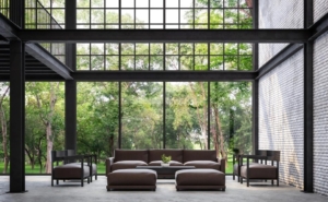 Spacious modern living area with floor-to-ceiling glass walls opening to a lush forest view, exemplifying biophilic design by blending indoor and outdoor environments.